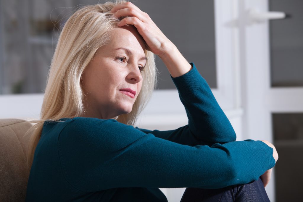 Sad depressed woman at home sitting on the couch, looking down and touching her forehead, loneliness and pain concept